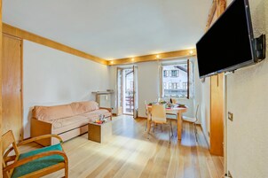 Una sala de estar con un sofá, una mesa de comedor con sillas, un televisor montado en la pared y un ventanal con vista a edificios.