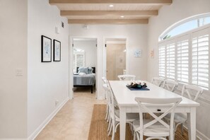 A bright, white interior with a dining area, a visible bedroom with a bed, and a large window with blinds.