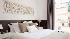 Ein ordentlich bezogenes Bett in Weiß und Beige, ein dekoratives Wandbild darüber und ein Blick auf ein Gebäude durch ein Fenster.