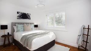 A bedroom with a bed, two bedside tables, a window with blinds, a wall-mounted picture, and a wooden rack with a towel.
