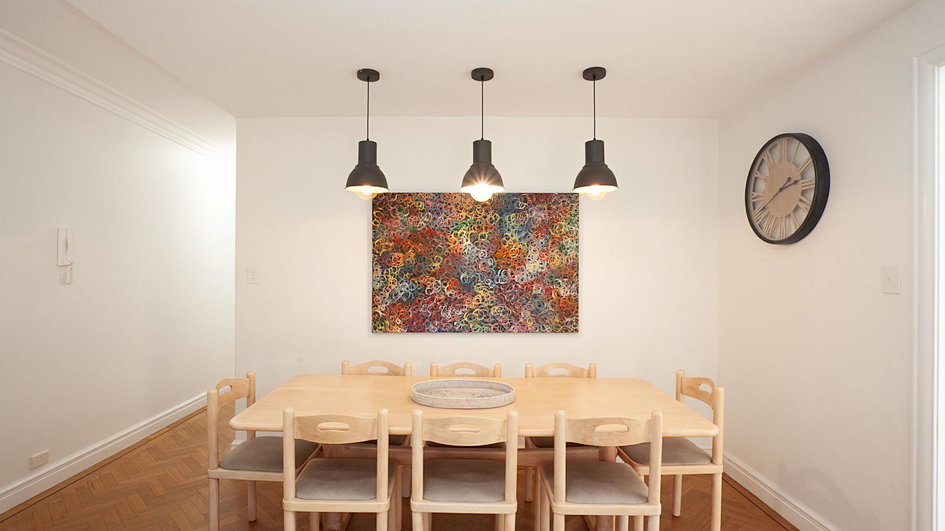 A dining area with a wooden table and chairs, three pendant lights, a colorful abstract painting, and a wall clock.