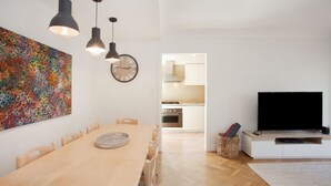 A modern living room with a wooden dining table, pendant lights, a large abstract painting, a wall clock, and a TV on a white cabinet.