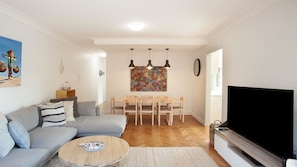 A living room with a grey sofa, a coffee table, a dining area with wooden chairs, a painting on the wall, and a flat-screen TV. - Scandi Bondi (Bondi Beach)