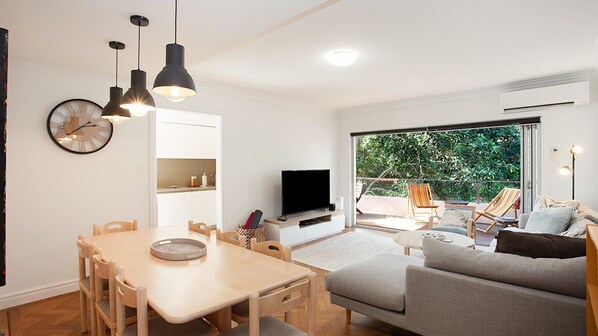 A modern living room with a dining area, a flat-screen TV, and a large sliding glass door leading to an outdoor patio. - Scandi Bondi (Bondi Beach)