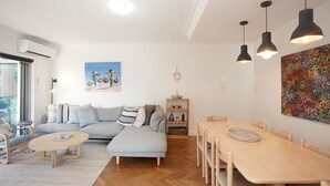 A modern living room with a grey sectional sofa, a wooden dining table with chairs, a coffee table, and pendant lighting. There is a painting on the wall and a colorful abstract piece above the table.