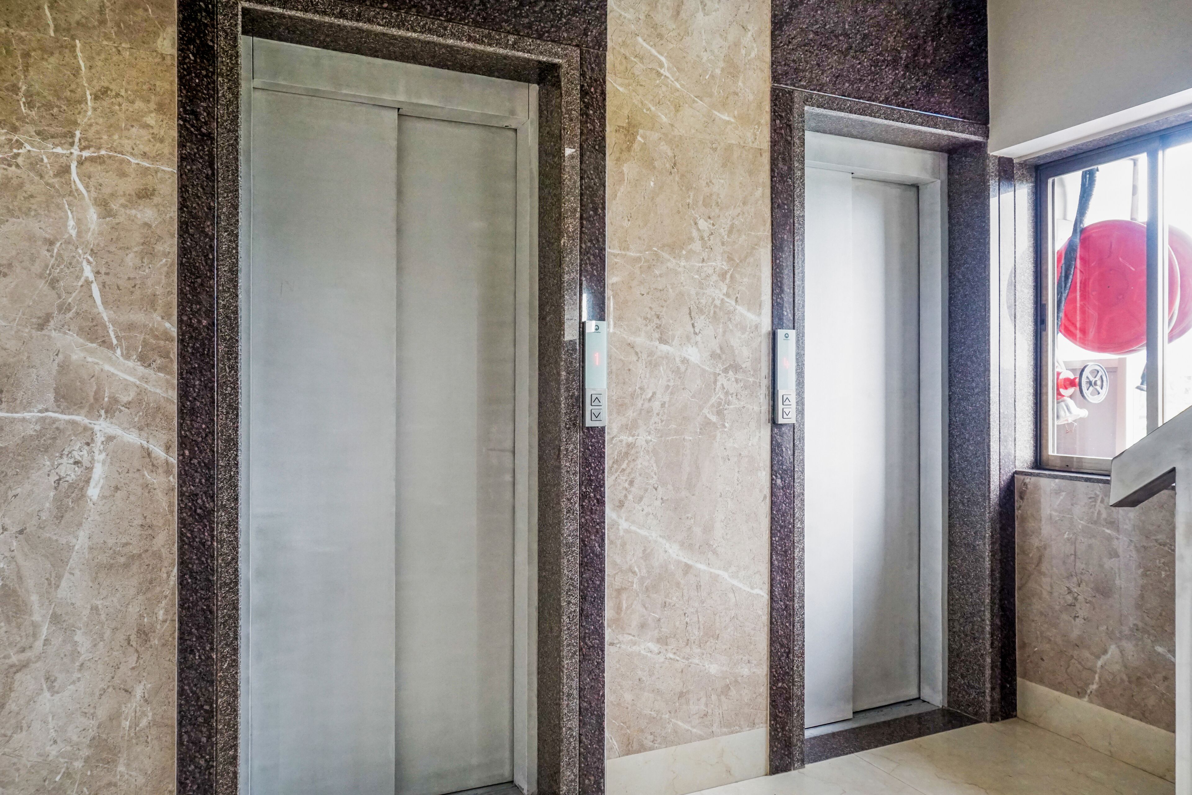 Two elevator doors with marble walls and a window with a red object outside.