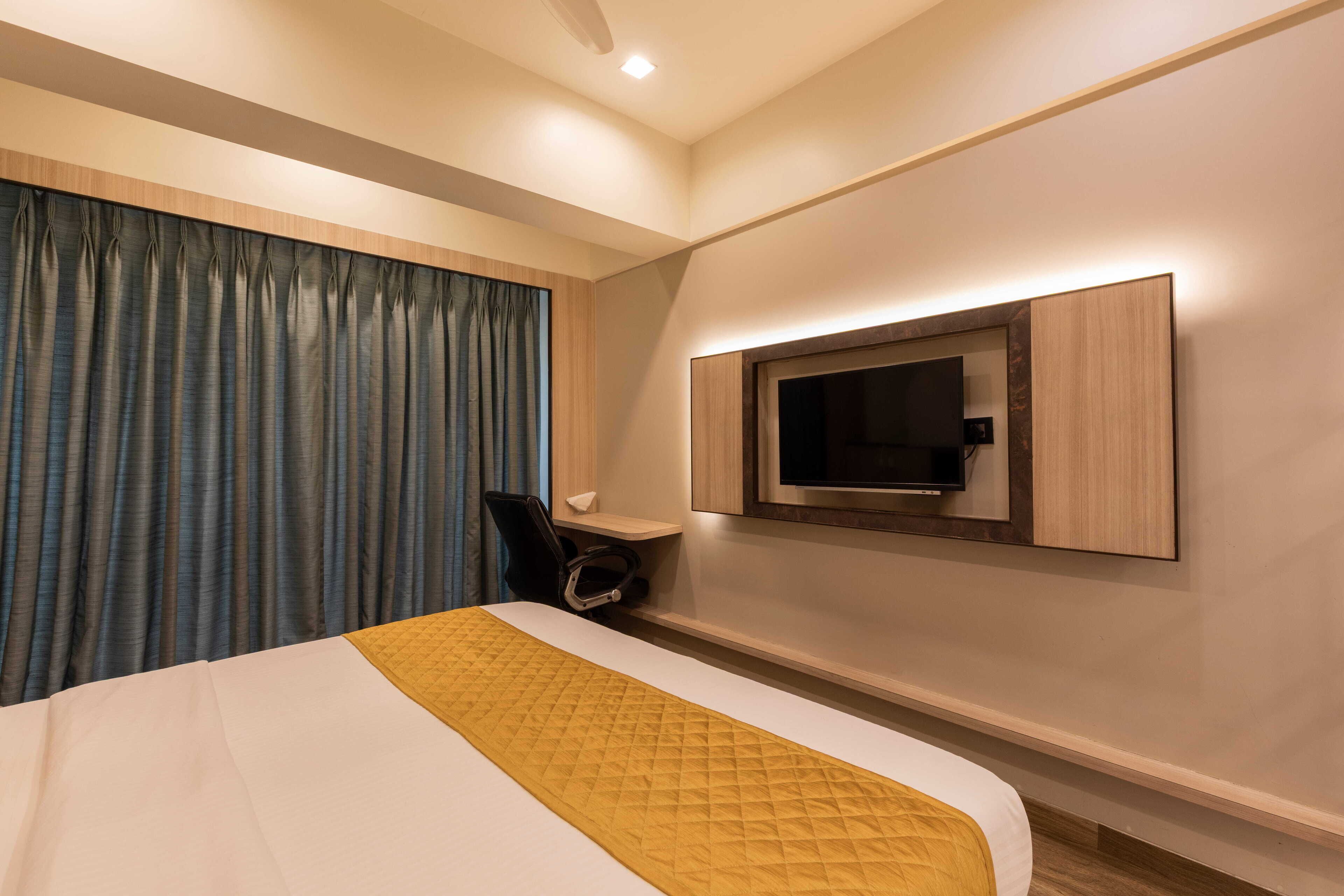 A hotel room with a bed, a desk, a chair, a TV mounted on the wall, and a window with curtains.