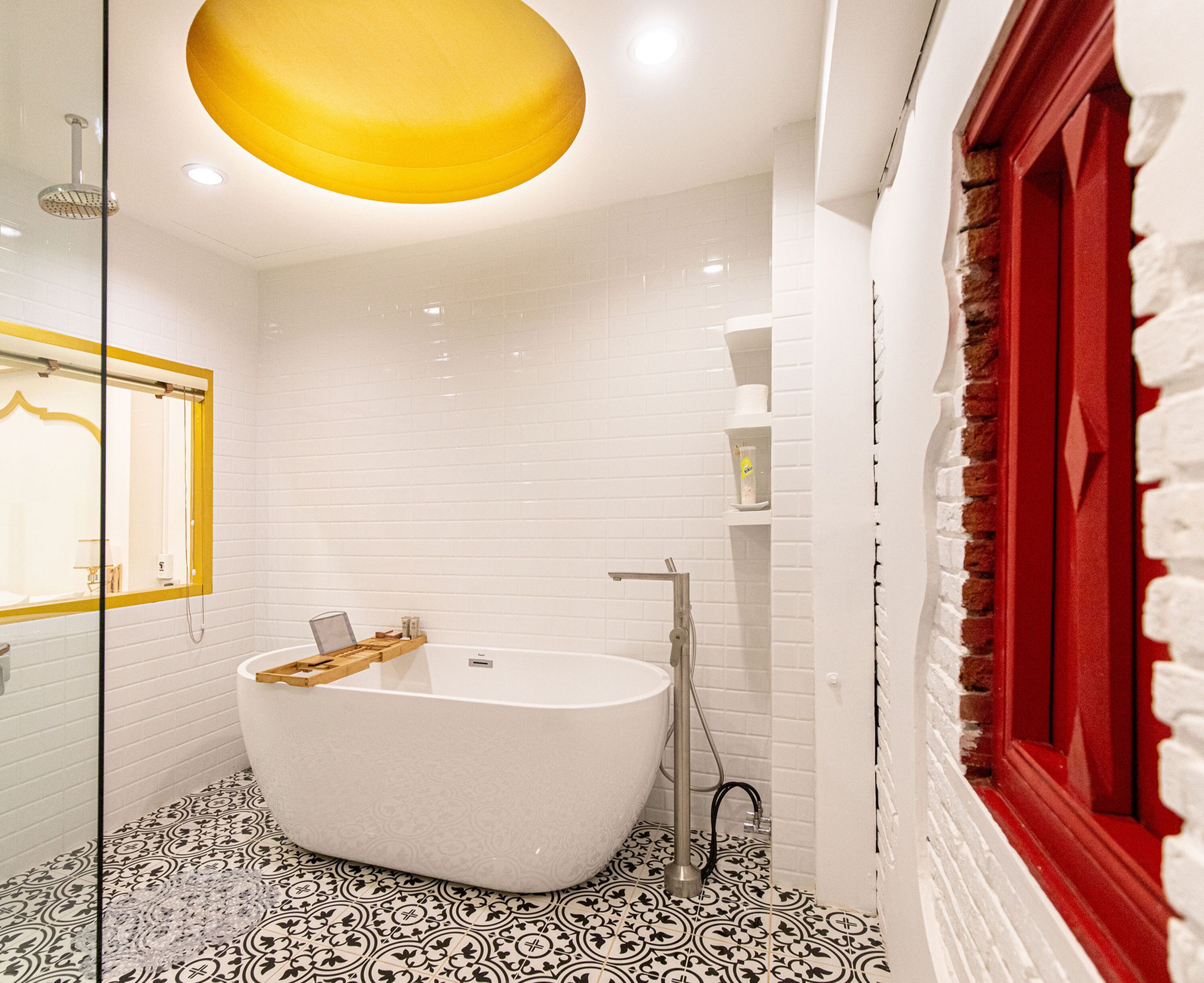 Un baño con una bañera independiente, una lámpara de luz amarilla, paredes de azulejos blancos y una ventana con marco rojo.