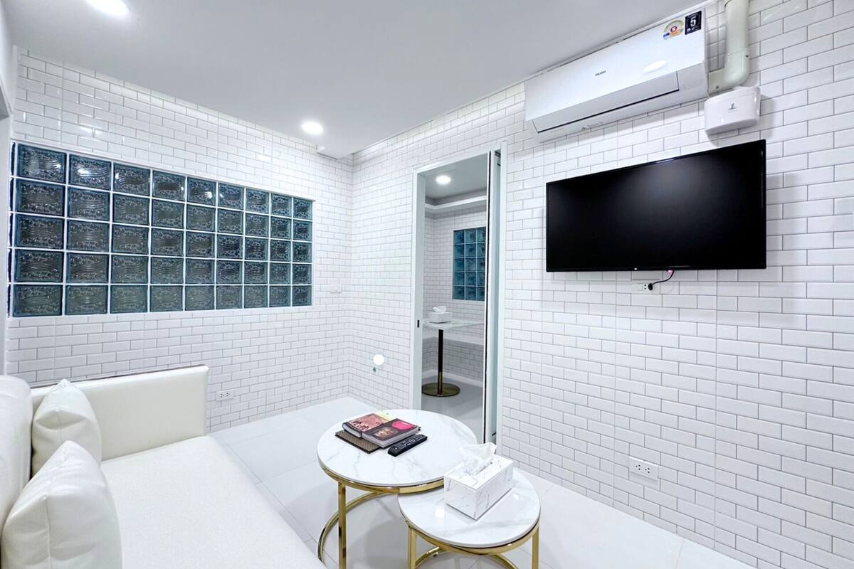 Une chambre moderne avec des murs recouverts de carreaux blancs, un téléviseur à écran plat fixé au mur, une porte en verre donnant sur une salle de bain, et une petite table ronde sur laquelle se trouvent un livre et des mouchoirs.