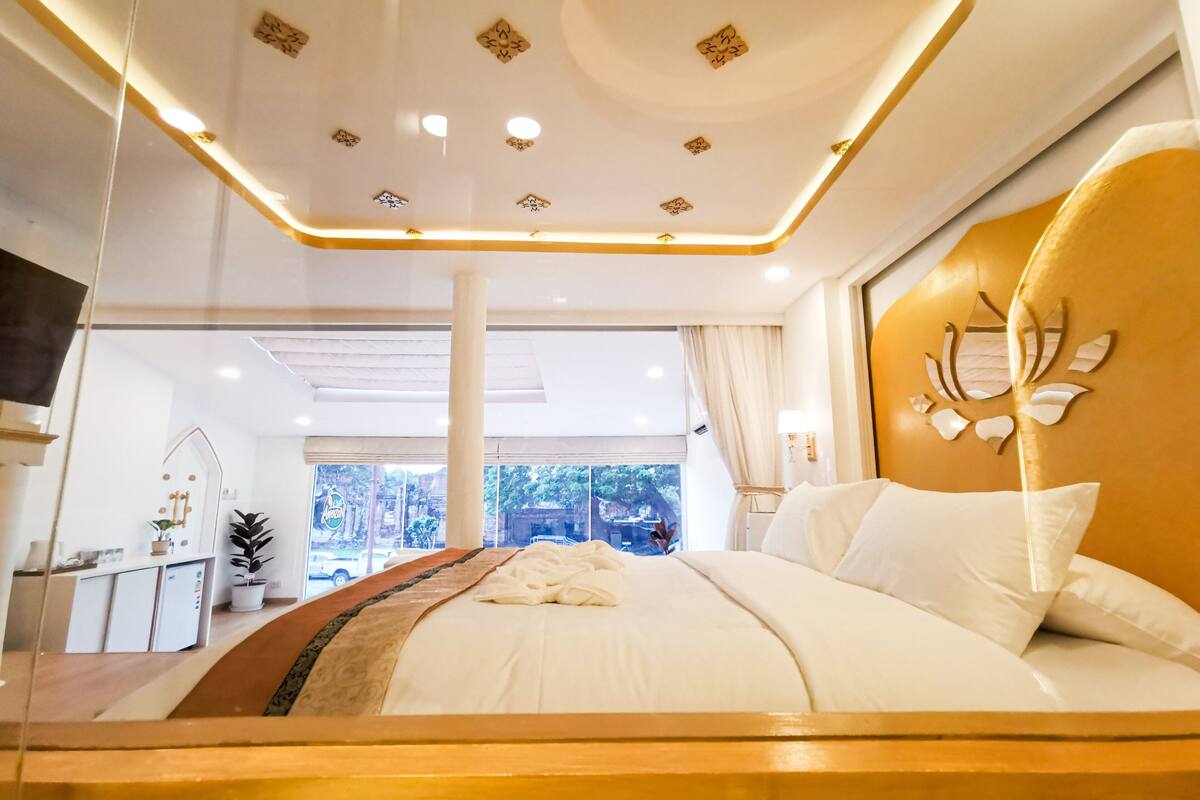 Une chambre moderne avec un grand lit, une tête de lit en bois, un plafond doté d’un éclairage encastré et une vue sur l’extérieur.
