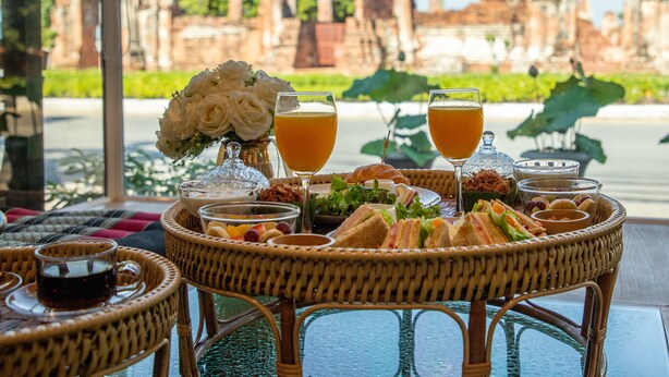 Una mesa dispuesta con diversas comidas y bebidas, como sándwiches, frutas y un vaso de zumo de naranja, con vistas a unas ruinas antiguas al fondo.