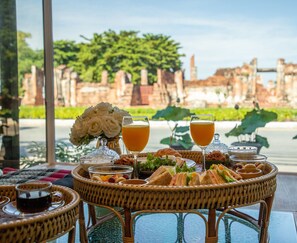 A table set with a variety of food and drinks, including sandwiches, fruits, and a glass of orange juice, with a view of ancient ruins in the background. - One Dhatu Ayutthaya Premium Homestay (Ayutthaya)