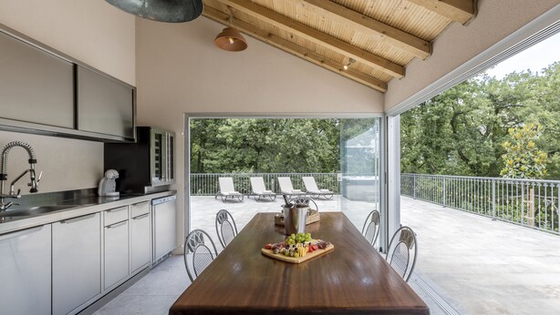 A modern kitchen with a wooden table set for dining, overlooking a balcony with outdoor seating.