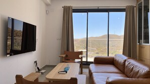 A modern living room with a large window, a flat-screen TV mounted on the wall, a wooden dining table, a leather sofa, and a small wooden chair.