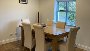 A dining area with a wooden table, four chairs, a hanging light fixture, a window with a view of greenery, and a framed picture on the wall.
