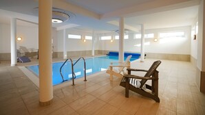 En indendørs swimmingpool med krystalklart vand, omgivet af hvide vægge og søjler, samt liggestole og solsenge.