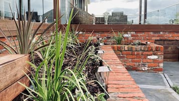 A rooftop garden with wooden planters, brick walls, and a glass railing overlooking a cityscape.