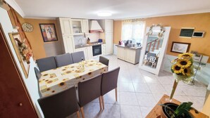 Een kleine keuken met een eethoek, met een tafel met een zonnebloem tafelkleed, een inbouwoven en een koelkast.
