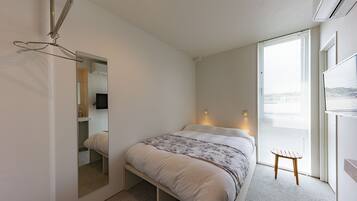 Un dormitorio con cama, un taburete de madera, un espejo y una ventana con persianas.