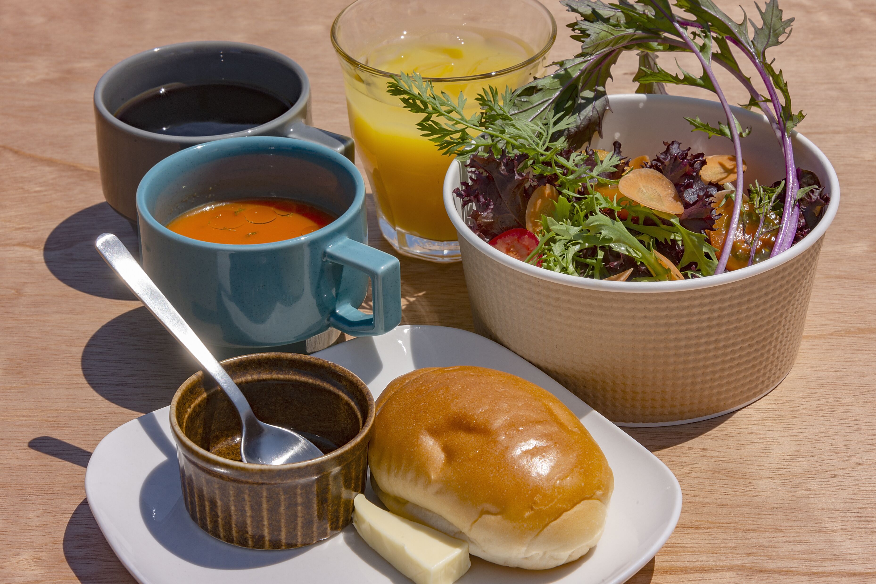 A meal consisting of soup, juice, salad, a bun, and a small bowl of sauce on a wooden table.