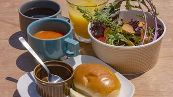A meal consisting of soup, juice, salad, a bun, and a small bowl of sauce on a wooden table.