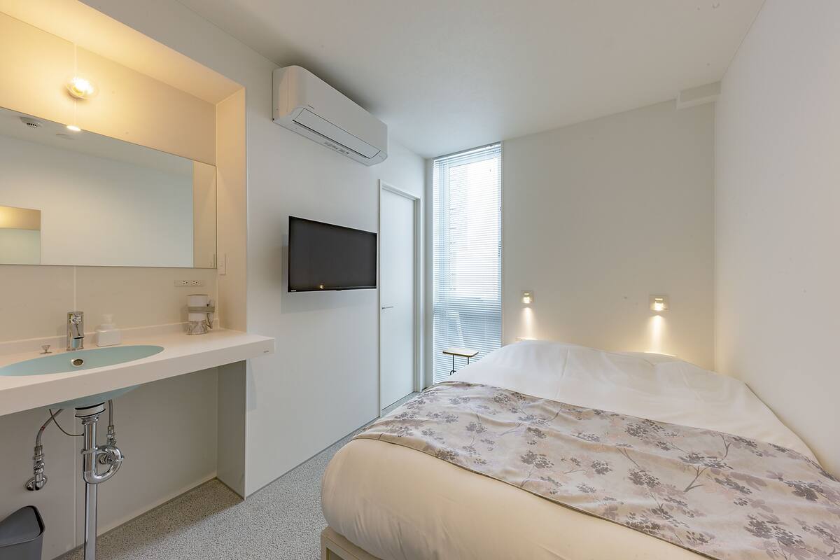 Ein Schlafzimmer mit einem Bett, einem großen Spiegel, einem an der Wand montierten Fernseher und einem Fenster mit Jalousien.