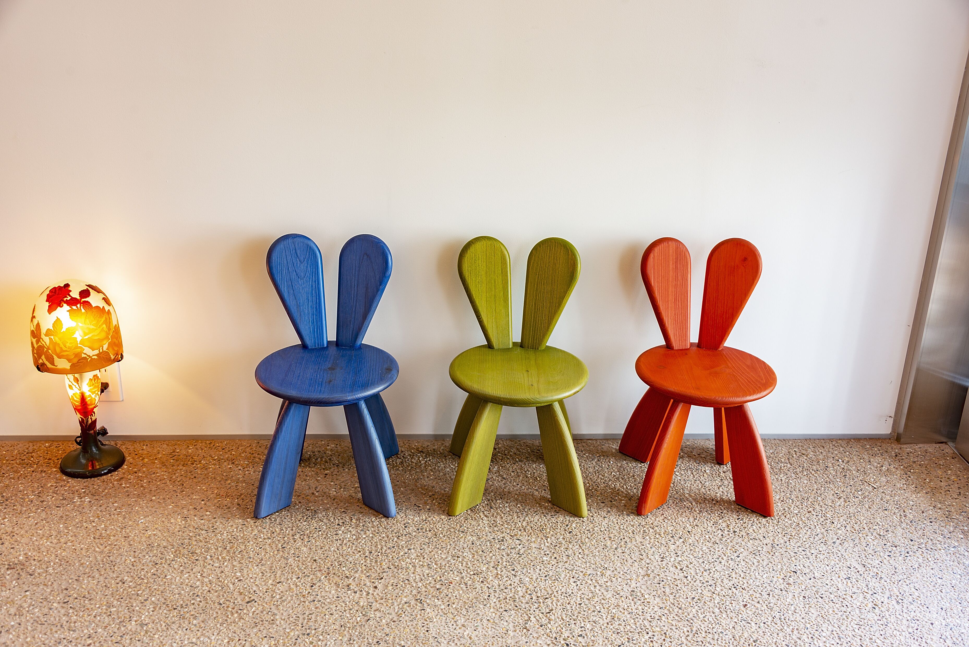 Three colorful chairs and a lamp against a plain wall.