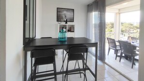 A dining area with a table and chairs, a pendant light, a framed picture on the wall, and a view of the outdoors through a glass door. - Cozy Air-Conditioned Apartment for 2 with Terrace, Near Beach, Fully Equipped Kitchen (Cap d'Agde naturisme)