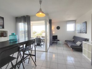 A modern living room with a sofa, a dining area with chairs, a TV, and a large window.