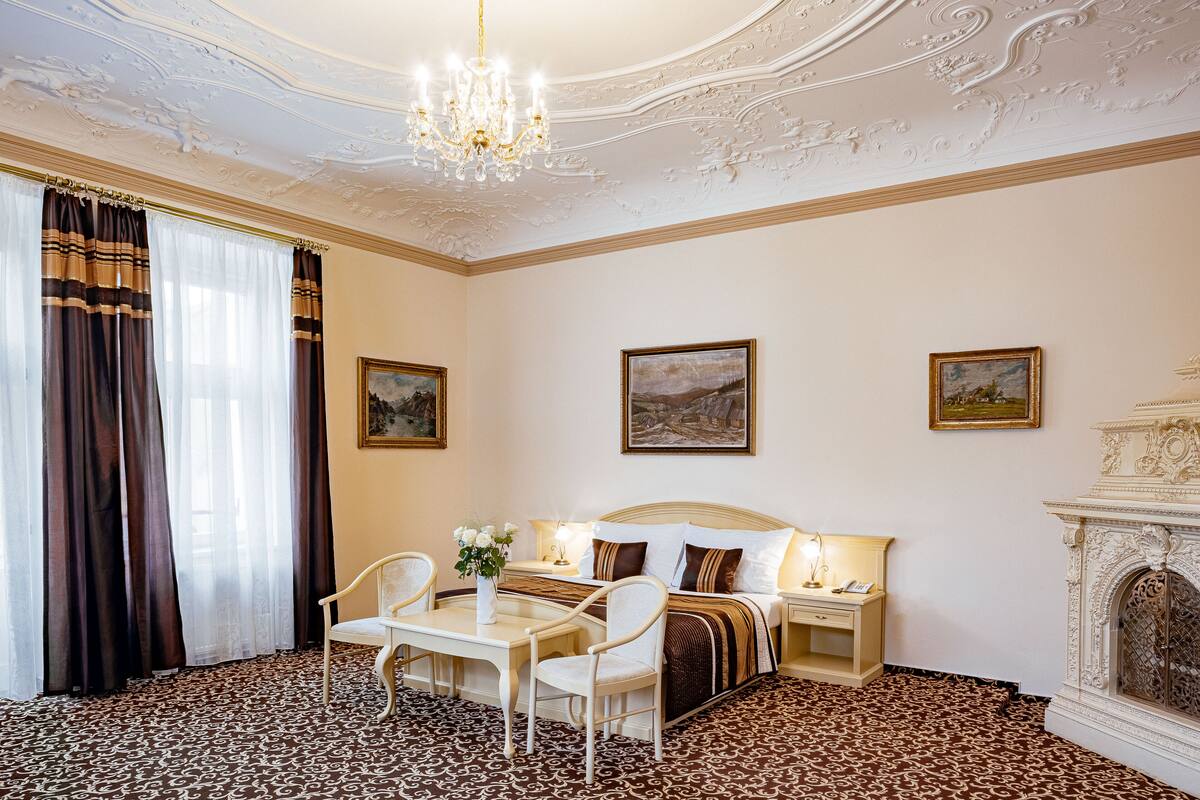A hotel room with a bed, two bedside tables, a chandelier, a fireplace, and a patterned carpet.