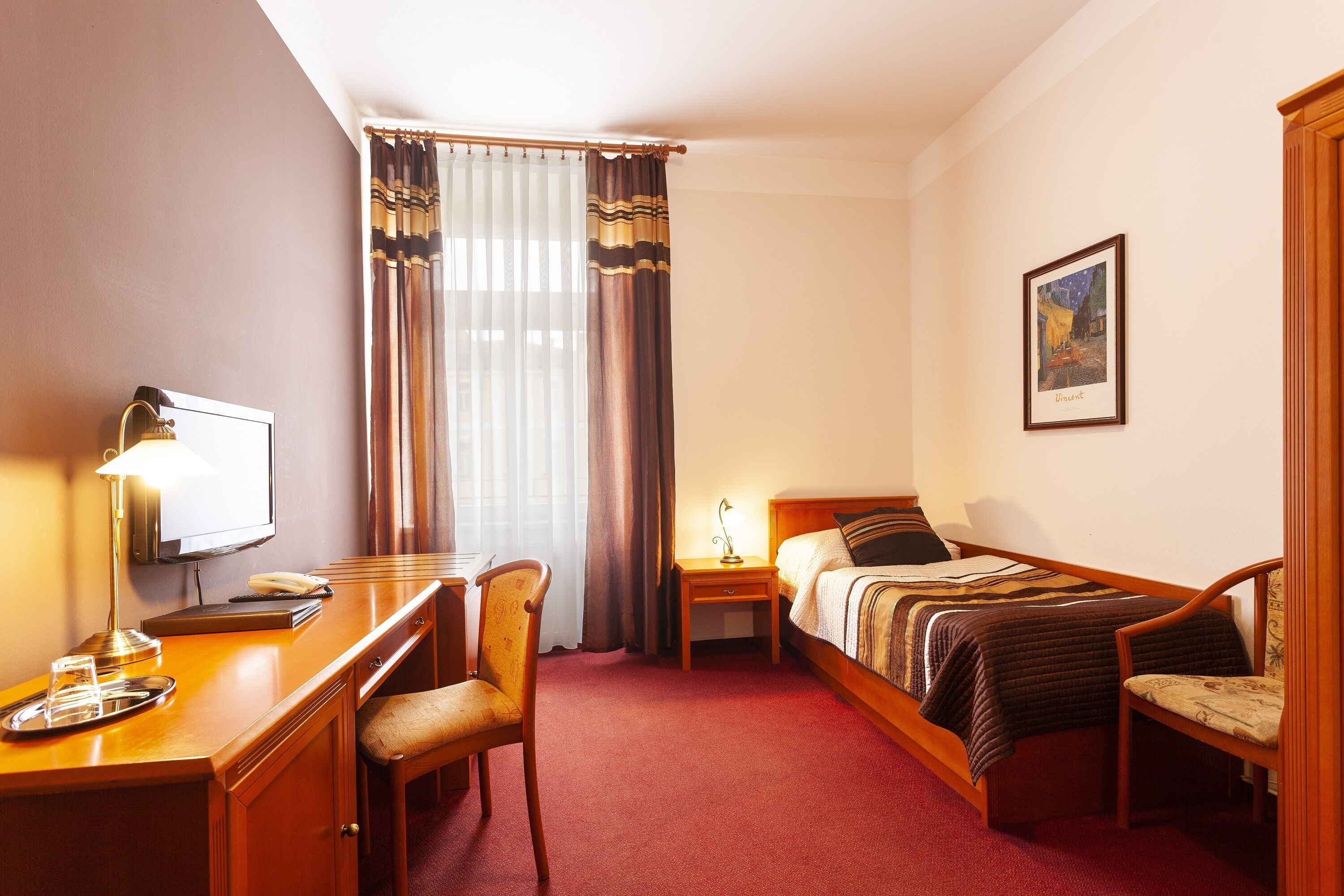 A hotel room with a bed, a desk with a lamp, a chair, a television, and a framed picture on the wall.