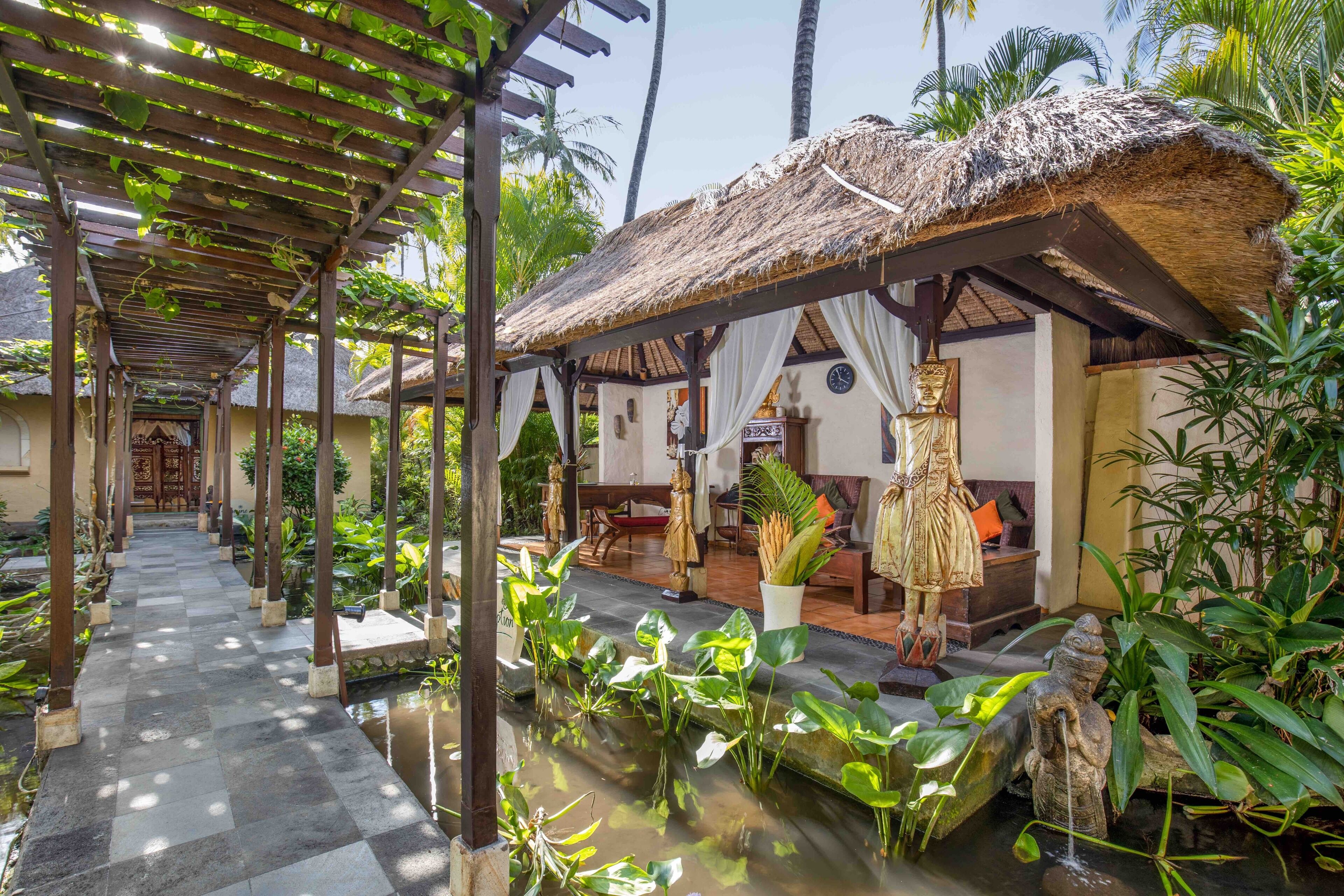 A covered walkway with a thatched roof, a small pond with water plants, and statues.