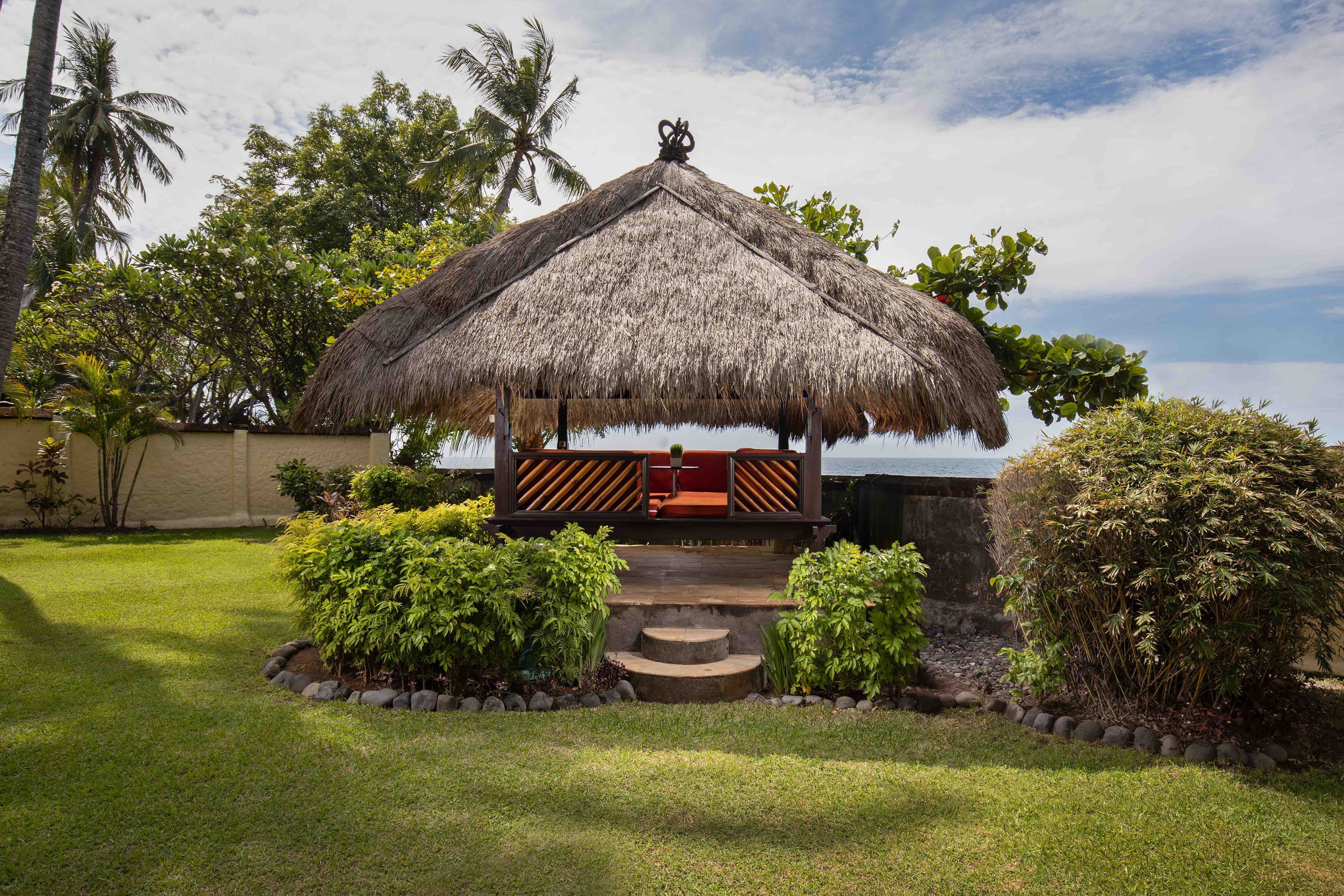 A thatched-roof structure with a wooden bench and steps leading up to it, surrounded by greenery and palm trees.