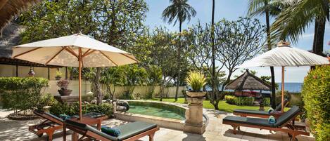 Zona de piscina con sillones reclinables y sombrillas, rodeada de vegetación tropical y vista al océano.