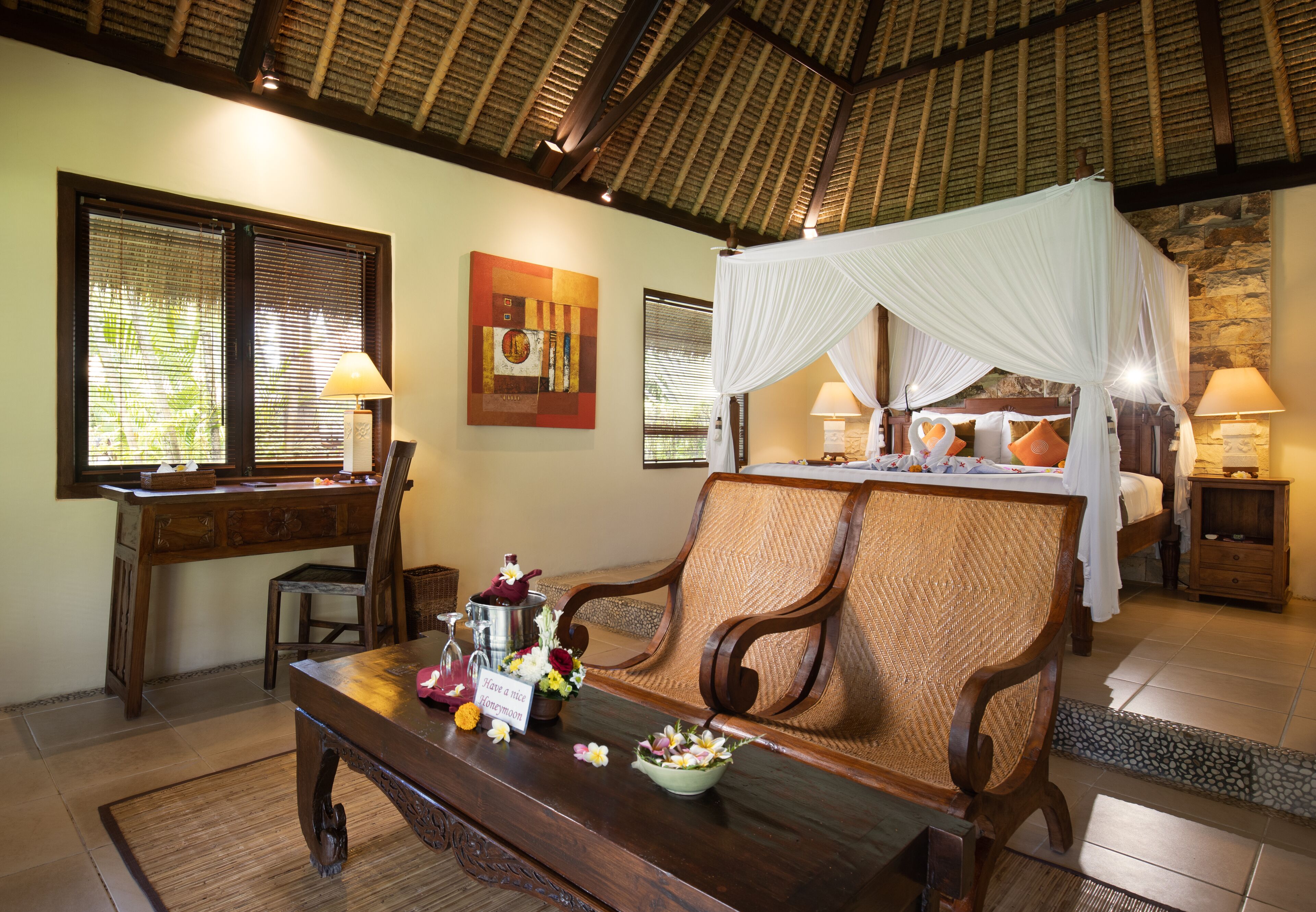 A room with a wooden desk, a cushioned bench, a bed with a canopy, and a stone wall.
