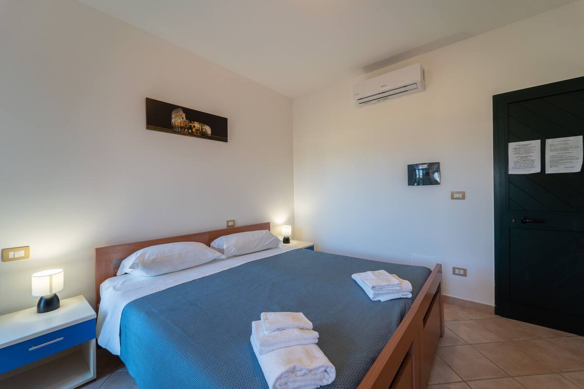 A neatly made bed with white linens and a blue bedspread, a bedside table with a lamp, a wall-mounted air conditioner, and a framed picture above the bed.