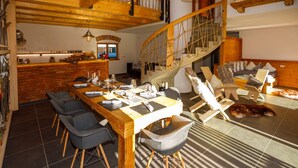A spacious dining area with a long wooden table set for a meal, surrounded by modern chairs. A staircase with a wooden railing leads to an upper level. - Alpenloft 1.0 (Mittenwald)