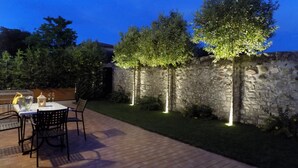 Een terras buiten, met een stenen muur, een tafel met stoelen en een kleine tuin met bomen en verlichting.