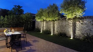 An outdoor patio with a stone wall, a table with chairs, and a small garden area with trees and lights.