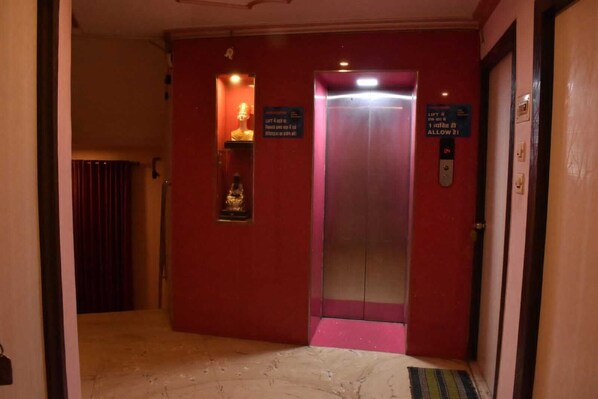 A hallway with a red wall, an elevator, and a small display case with a statue. - Hotel Shri Ram International (Varanasi)