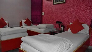 A hotel room with three beds, each with white bedding and red pillows. There is a small table with a lamp and a coffee maker. - Hotel Shri Ram International (Varanasi)
