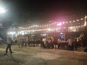 An outdoor dining area with tables and chairs, a stage with a sign, and a crowd of people.