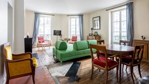 A room with a green sofa, wooden dining table, and chairs, a red armchair, and a small cabinet. - Picture Postcard, by Veeve (Paris)