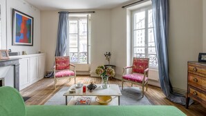 A room with a fireplace, two chairs, a table with a vase of flowers, and a window with curtains. - Picture Postcard, by Veeve (Paris)