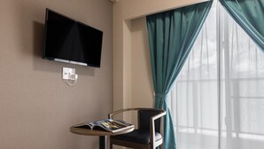 A hotel room with a flat-screen TV mounted on the wall, a small round table with a magazine, a chair, and a window with teal curtains. - Mr.KINJO LINK in Kugai Miyakojima (Miyakojima)