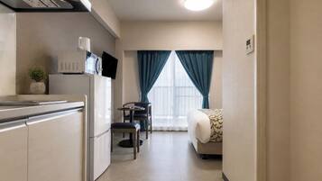 A compact hotel room with a kitchenette, a small dining table, a single bed, and a large window with curtains.