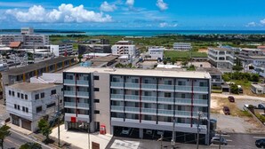 A modern multi-story building with a flat roof, surrounded by other buildings and a clear view of the ocean. - Mr.KINJO LINK in Kugai Miyakojima (Miyakojima)