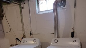 Two white front-loading washing machines in a utility room with exposed pipes and a small window.