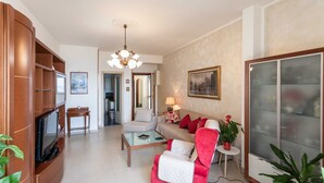 Ein Wohnzimmer mit einer Couch, Sesseln, einem Couchtisch, einem Fernseher, einer Chandelüster und einer Vitrine.