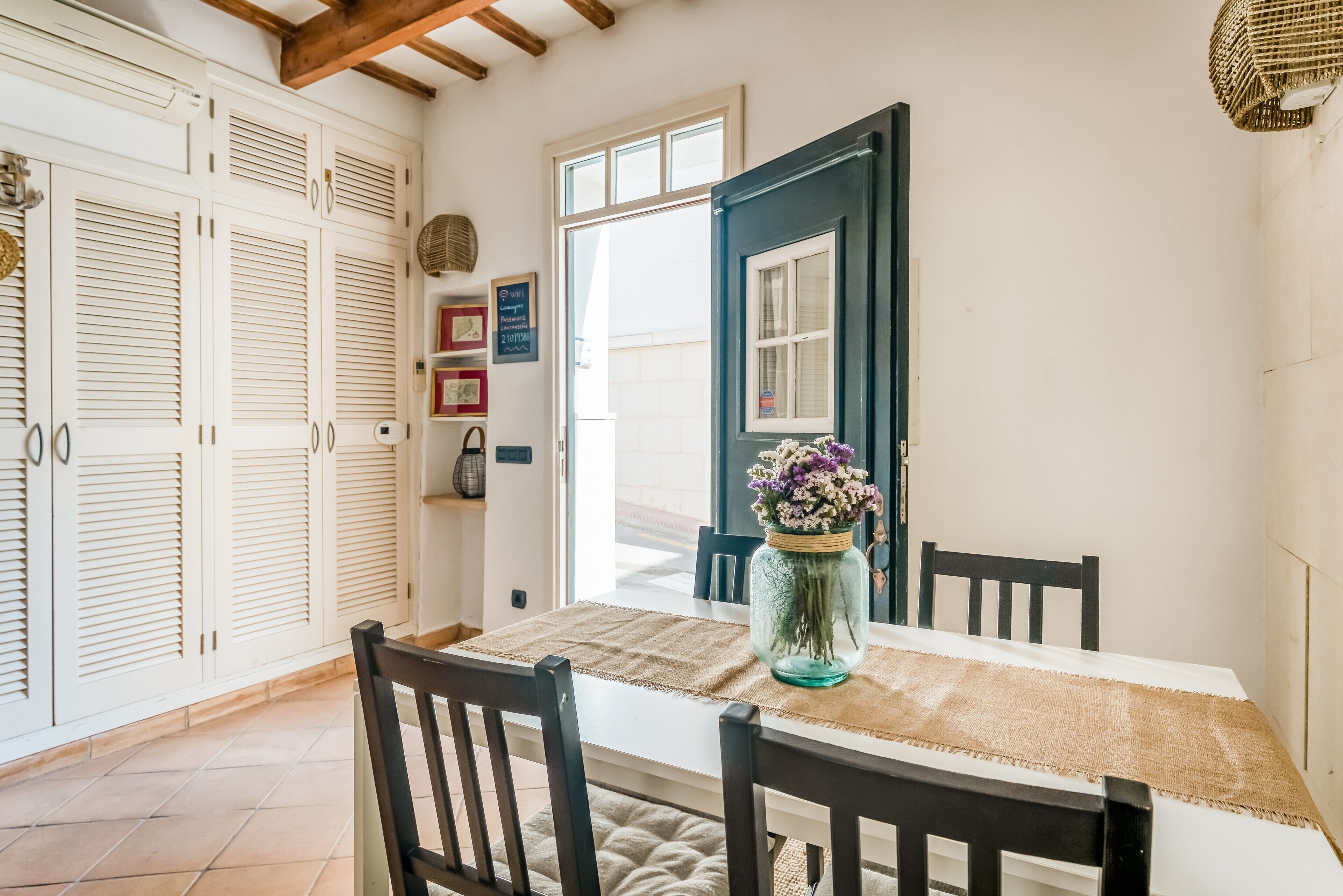 A dining area with a table set for two, a vase with flowers, and a view of the outside.
